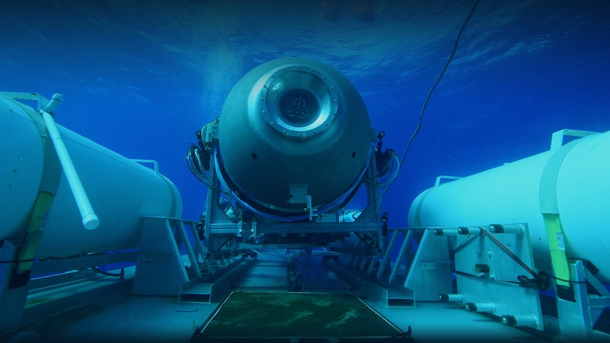 A submersible vessel is seen underwater, attached to a larger platform