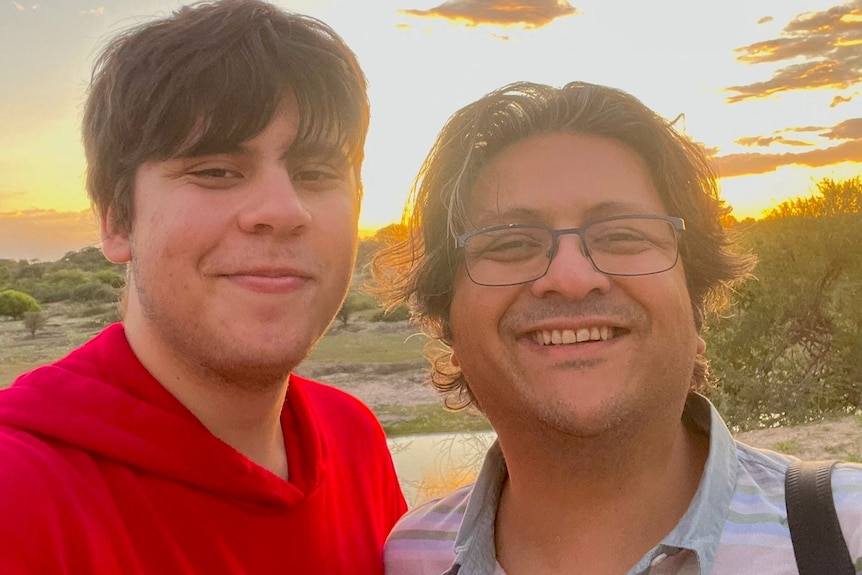 A young man on the left wearing a red hoodie, next to him an older man with grey-ish hair, wearing glasses. Both are smiling.