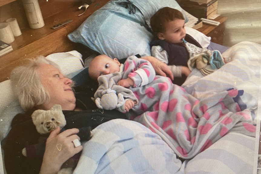 An older woman lays in bed with two small children, all cuddling their teddies.